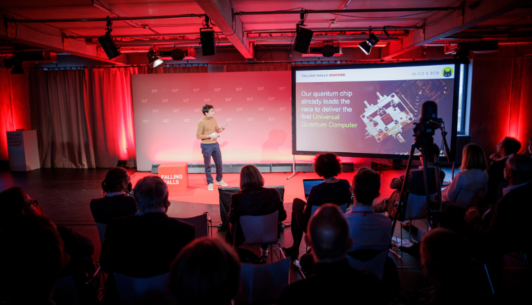 a contestant gives their venture pitch presentation to an audience on a large projection screen, the back wall lit red